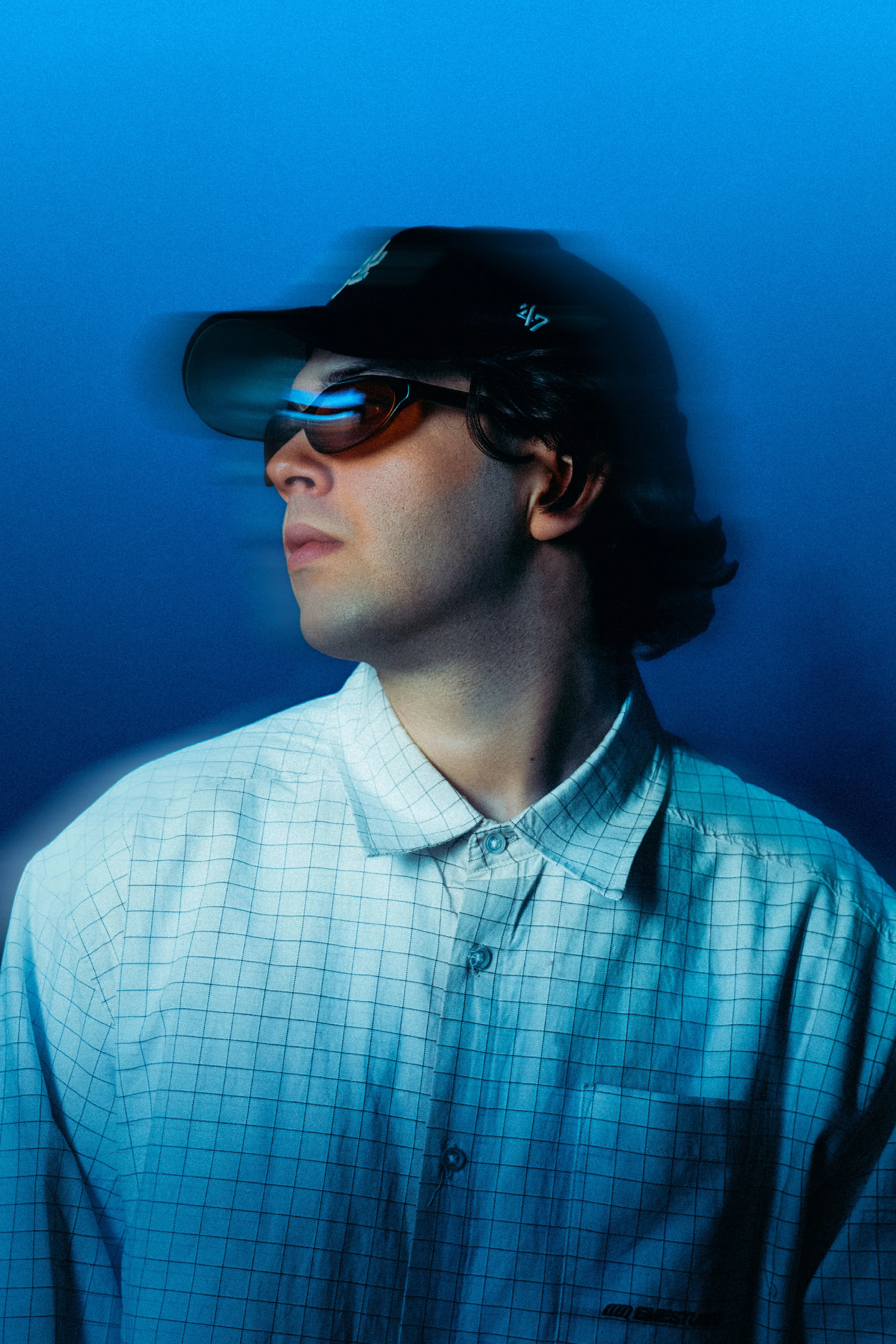 AMANN in a checked shirt and cap against a blue backdrop