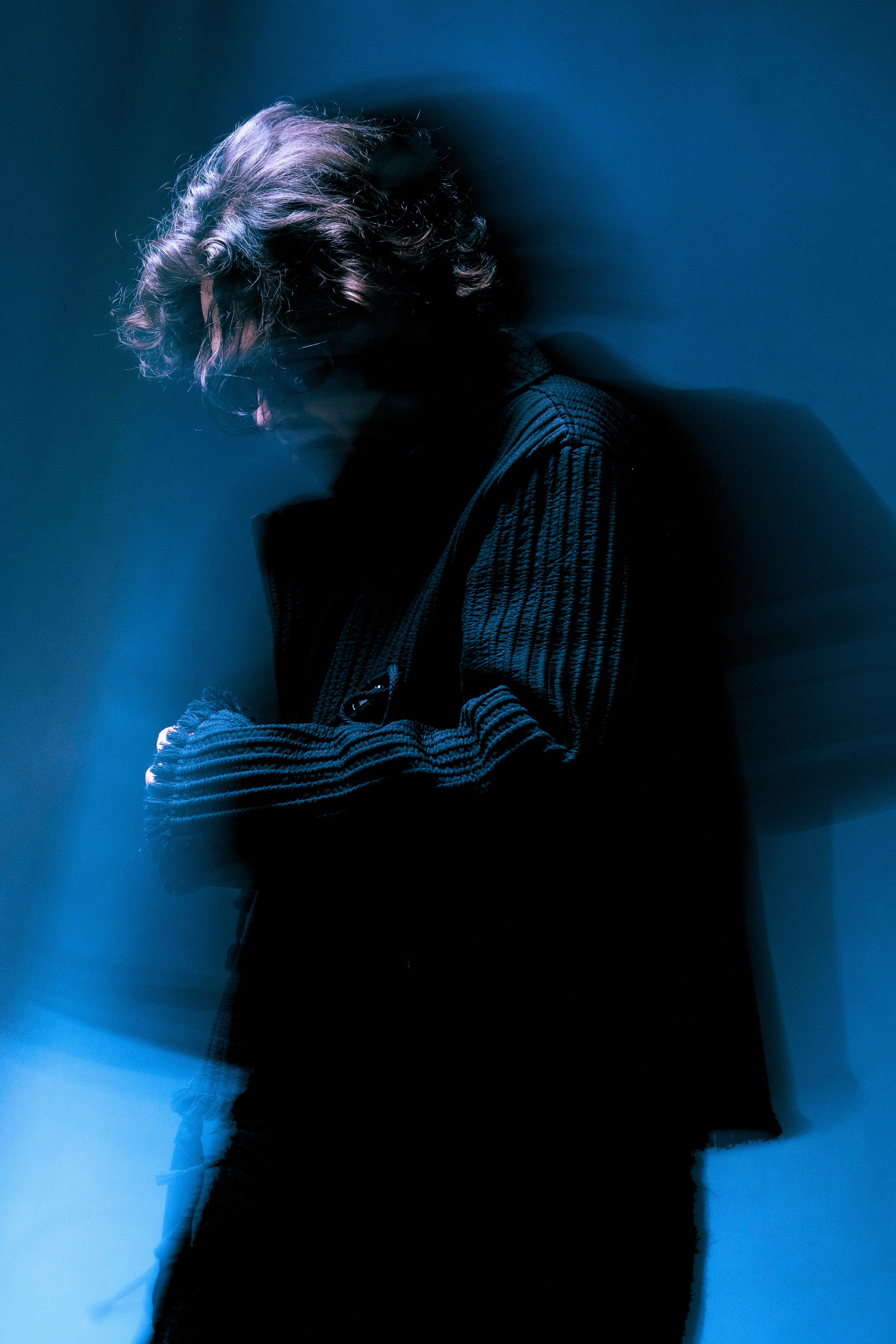 AMANN looking down, motion-blurred, in a ribbed black jacket against a blue backdrop